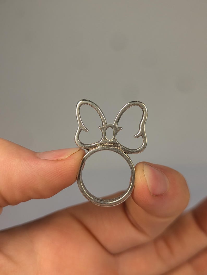 May include: A silver-toned metal ring featuring a cutout bow design on the top. The center of the bow includes a small, oval-shaped opening. The word Disney is stamped in small, clear letters at the base of the bow where it meets the ring band.