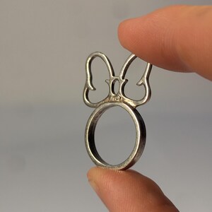 May include: A silver-toned metal ring featuring a decorative top element shaped like a stylized bow or ears. The metal band is circular, and the piece includes a small, stamped hallmark visible at the base of the bow design on the front surface.