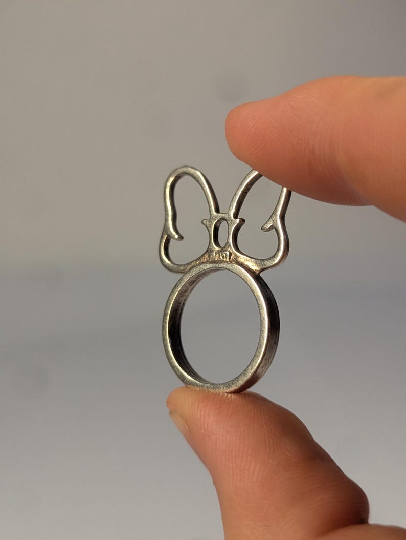 May include: A silver-toned metal ring featuring a decorative top element shaped like a stylized bow or ears. The metal band is circular, and the piece includes a small, stamped hallmark visible at the base of the bow design on the front surface.