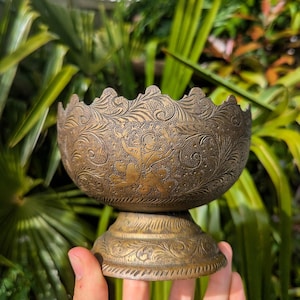 May include: A round brass bowl with a pedestal base, featuring intricate etched floral patterns and a central figure engraving. The metal surface has a dark, aged patina with a scalloped rim edge and detailed scrollwork covering the entire exterior.