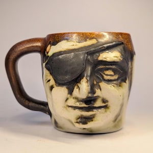 May include: A ceramic mug shaped like a human face with a prominent eye patch over the left eye. The mug features a glossy brown rim and handle, with a cream-colored base and dark, textured glaze accents highlighting the facial features and eye patch.