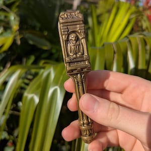 May include: A vintage brass-colored nutcracker with a detailed design. The top section features an embossed image of a seated figure and the dates 1562-1616. The handle has a decorative, fluted design.