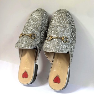 Puede incluir: Un par de mocasines sin cordones con purpurina plateada y un detalle de bocado de metal dorado en el empeine. La plantilla interior es de color beige claro con un parche rojo en forma de corazón en el talón y el nombre Gucci en dorado.
