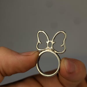 May include: A silver-toned metal ring featuring a cutout bow design on the top. The center of the bow includes a small, oval-shaped detail with the word Disney engraved in a clear, legible font on the band section just below the bow.