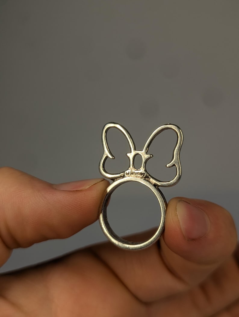 May include: A silver-toned metal ring featuring a cutout bow design on the top. The center of the bow includes a small, oval-shaped detail with the word Disney engraved in a clear, legible font on the band section just below the bow.