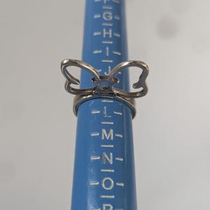 May include: A silver-toned metal ring featuring a bow-shaped design element on the top. The ring is positioned on a blue plastic ring sizing mandrel that displays alphabetical size markings, with the ring resting between the K and L size indicators.