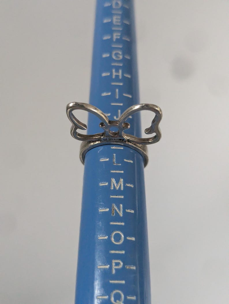 May include: A silver-toned metal ring featuring a bow-shaped design element on the top. The ring is positioned on a blue plastic ring sizing mandrel that displays alphabetical size markings, with the ring resting between the K and L size indicators.