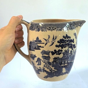 Può includere: Una brocca in ceramica con base color crema e un motivo decorativo blu scuro. Il design raffigura un paesaggio con pagode, alberi di salice e due uccelli in volo. Presenta un manico curvo e un beccuccio sul bordo superiore.