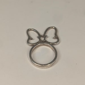 May include: A silver-toned metal ring featuring a decorative bow-shaped element attached to the top of the circular band. The bow design consists of two symmetrical loops with a small oval center piece, creating a distinct silhouette on the finger.