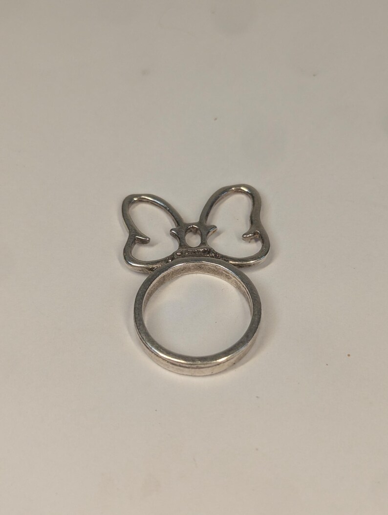 May include: A silver-toned metal ring featuring a decorative bow-shaped element attached to the top of the circular band. The bow design consists of two symmetrical loops with a small oval center piece, creating a distinct silhouette on the finger.