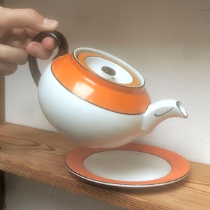 May include: A white ceramic teapot with a bright orange band around the top and a dark brown handle. The teapot features a matching lid with a small brown knob and a white spout. A matching white saucer with an orange rim sits underneath.