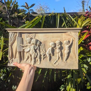 May include: A rectangular, beige relief sculpture showing several cherubic figures in different poses. One figure paints at an easel, while others interact, suggesting a playful scene. The artwork is held by a hand.