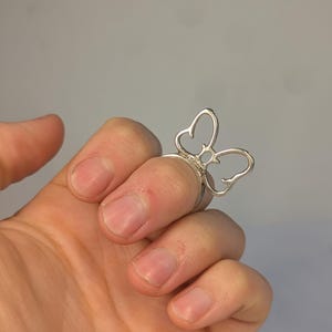 May include: A silver-toned ring featuring a butterfly-shaped design element on the front. The butterfly is constructed from thin, curved metal wire forming an open, outline-style shape that sits atop the finger band. The metal has a smooth finish.