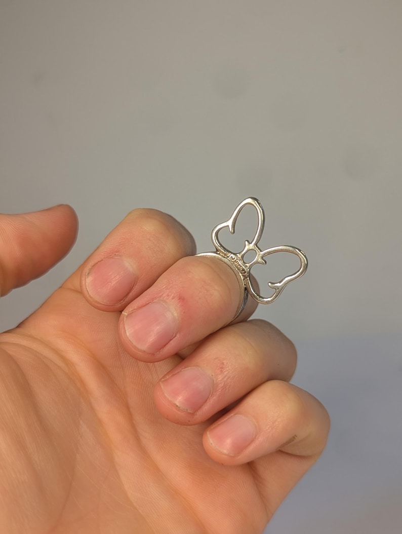 May include: A silver-toned ring featuring a butterfly-shaped design element on the front. The butterfly is constructed from thin, curved metal wire forming an open, outline-style shape that sits atop the finger band. The metal has a smooth finish.