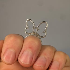 May include: A silver-toned ring featuring a stylized, open-work bow design. The bow is positioned vertically on the band, with thin, curved metal lines forming the shape of the bow. The metal has a polished finish and a small hallmark is visible.
