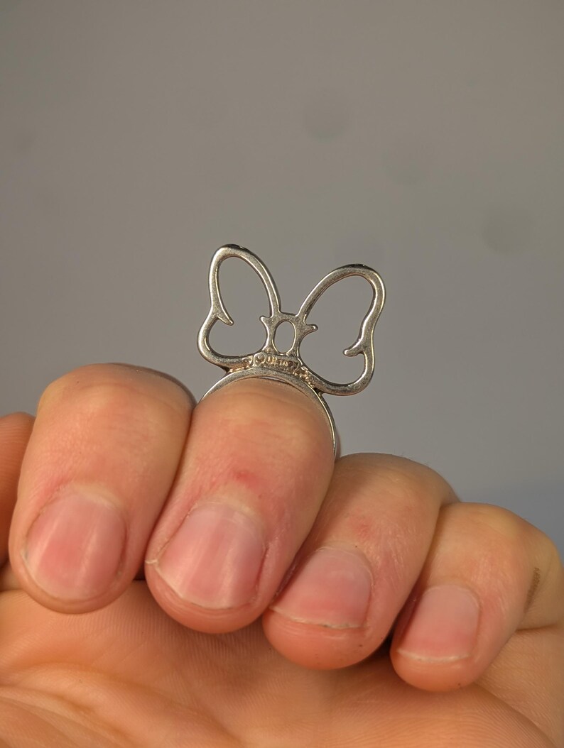 May include: A silver-toned ring featuring a stylized, open-work bow design. The bow is positioned vertically on the band, with thin, curved metal lines forming the shape of the bow. The metal has a polished finish and a small hallmark is visible.