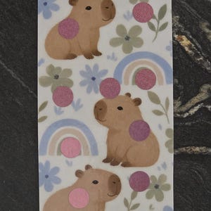 May include: A rectangular sticker sheet featuring three brown capybara illustrations, pastel blue rainbows, and muted green and blue flowers. The design includes pink circular accents, the text Lee-Arne's Creations for you, Save $30, and Denomination $1-$6.