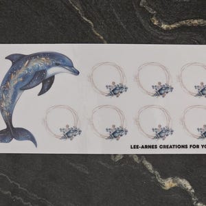 May include: A rectangular sticker sheet featuring a large blue dolphin illustration with gold accents on the left side and seven smaller circular frames decorated with blue seashells on the right. The text LEE-ARNES CREATIONS FOR YOU is printed in black.