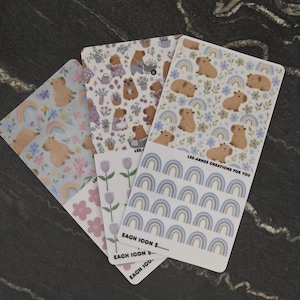 May include: Three rectangular sticker sheets featuring capybara illustrations, floral patterns, and rainbow motifs. The sheets display various capybara poses, small flowers, and rows of rainbow icons. Text on the sheets includes LEE-ARNES CREATIONS FOR YOU.