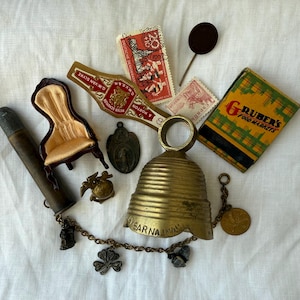 May include: A collection of vintage items including a brass bell with a chain and charms, a miniature chair with gold fabric, a cigar band, two postage stamps, a matchbox labeled Gruber's Food Markets, a metal pin, and a cylindrical metal tube.