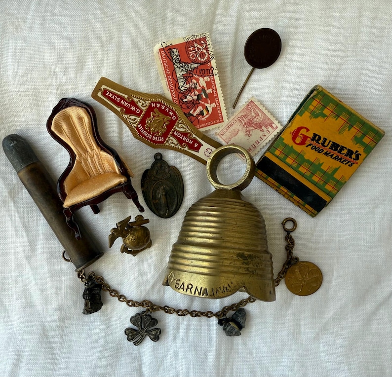May include: A collection of vintage items including a brass bell with a chain and charms, a miniature chair with gold fabric, a cigar band, two postage stamps, a matchbox labeled Gruber's Food Markets, a metal pin, and a cylindrical metal tube.