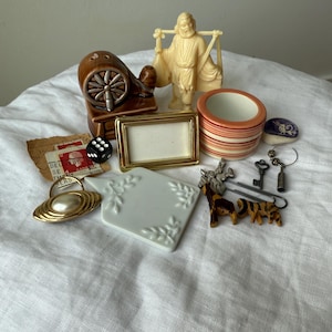 May include: A collection of small objects including a brown ceramic spinning wheel, a cream-colored figurine of a person carrying a yoke, a gold-toned picture frame, a striped ceramic cup, a black die, a white ceramic tile, and various metal trinkets.