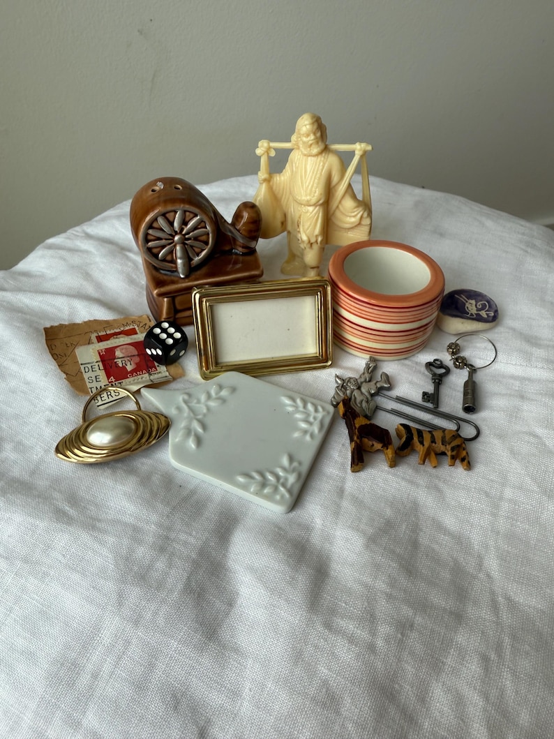 May include: A collection of small objects including a brown ceramic spinning wheel, a cream-colored figurine of a person carrying a yoke, a gold-toned picture frame, a striped ceramic cup, a black die, a white ceramic tile, and various metal trinkets.