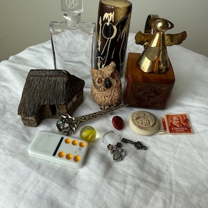 May include: A collection of vintage items including a clear glass decanter, a brown and black cylindrical container, a brass angel figurine on a wooden block, a small house model, an owl figurine, a metal pin, a domino, a marble, a key, and a stamp.