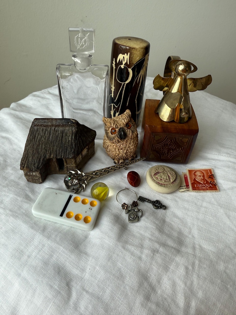 May include: A collection of vintage items including a clear glass decanter, a brown and black cylindrical container, a brass angel figurine on a wooden block, a small house model, an owl figurine, a metal pin, a domino, a marble, a key, and a stamp.
