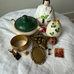 May include: A collection of vintage items including a ceramic Japanese doll, a green and cream lidded bowl, a brass bangle, a star-shaped face ornament, a brown button, a gold-tone oval frame, a small pin, and a red Spanish postage stamp.