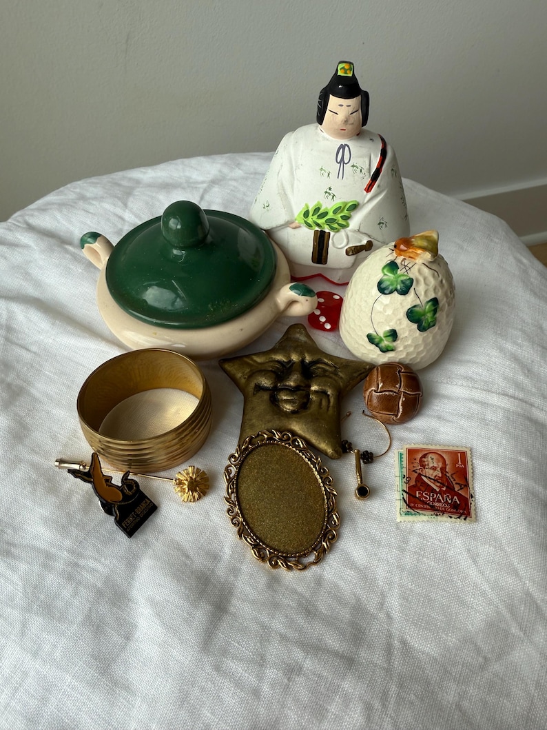 May include: A collection of vintage items including a ceramic Japanese doll, a green and cream lidded bowl, a brass bangle, a star-shaped face ornament, a brown button, a gold-tone oval frame, a small pin, and a red Spanish postage stamp.