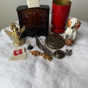 May include: A collection of small decorative items including a dark brown glass piano, a red glass vessel, a ceramic dog figurine, a brass angel bell, a metal feather pin, a circular bronze medallion, a postage stamp, and miniature animal carvings.