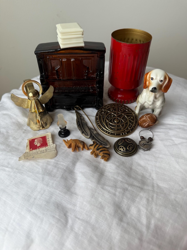 May include: A collection of small decorative items including a dark brown glass piano, a red glass vessel, a ceramic dog figurine, a brass angel bell, a metal feather pin, a circular bronze medallion, a postage stamp, and miniature animal carvings.