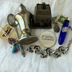 May include: A collection of vintage items including a silver-toned shaker, a metal house-shaped object, a silver spoon, a blue glass perfume bottle, a beaded figure, a round 1st edition token, a religious pendant, stamps, and a floral rhinestone bracelet.
