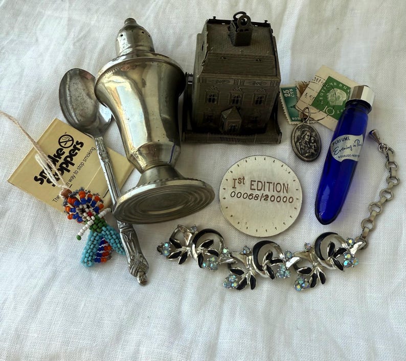 May include: A collection of vintage items including a silver-toned shaker, a metal house-shaped object, a silver spoon, a blue glass perfume bottle, a beaded figure, a round 1st edition token, a religious pendant, stamps, and a floral rhinestone bracelet.