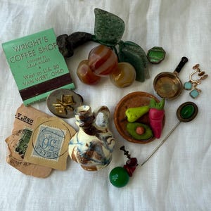 May include: A collection of vintage items including a green matchbook from Wright's Coffee Shop, a stone fruit brooch, a small ceramic pitcher, a miniature skillet, a stick pin with a green stone, a stamp, and various small decorative charms.