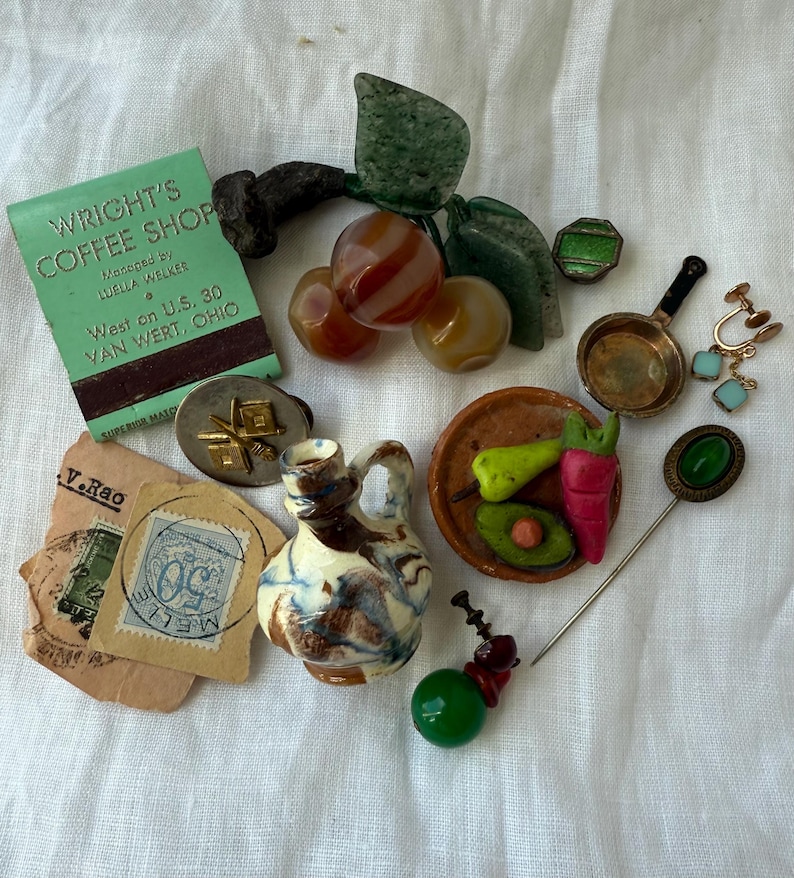 May include: A collection of vintage items including a green matchbook from Wright's Coffee Shop, a stone fruit brooch, a small ceramic pitcher, a miniature skillet, a stick pin with a green stone, a stamp, and various small decorative charms.