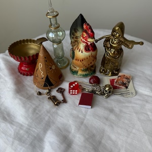 May include: A collection of small decorative objects including a red scalloped bowl, a clear glass bottle, a ceramic rooster figurine, a brass figure, a conical wooden piece with drawings, a red die, a skeleton charm, a key, and a miniature book.