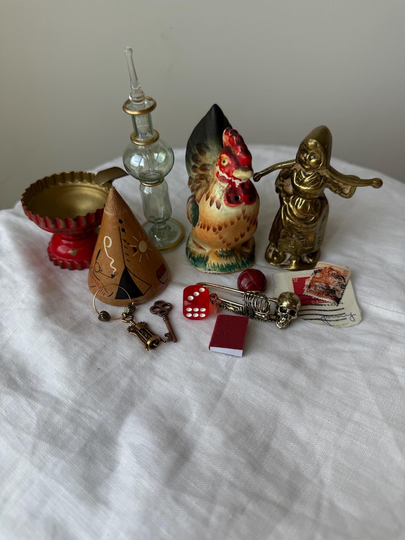 May include: A collection of small decorative objects including a red scalloped bowl, a clear glass bottle, a ceramic rooster figurine, a brass figure, a conical wooden piece with drawings, a red die, a skeleton charm, a key, and a miniature book.