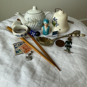May include: A collection of vintage items including a white ceramic teapot with blue floral patterns, a bell with blue flowers, two wooden sticks, a silver spoon with charms, a small figurine, a napkin ring, dice, stamps, and a copper coin.