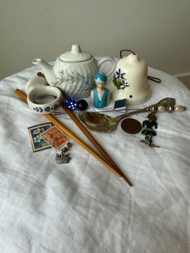 May include: A collection of vintage items including a white ceramic teapot with blue floral patterns, a bell with blue flowers, two wooden sticks, a silver spoon with charms, a small figurine, a napkin ring, dice, stamps, and a copper coin.