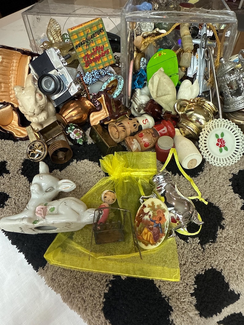May include: A collection of assorted vintage trinkets including a miniature camera, a green box labeled Headquarters for Thrifty Shoppers, a ceramic deer, a yellow organza bag, a silver pig figurine, and various small decorative metal and ceramic items.