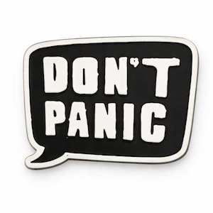 Puede incluir: Pin esmaltado en blanco y negro con forma de bocadillo. El pin presenta las palabras "DON'T PANIC" en letras blancas en negrita sobre un fondo negro, con un borde blanco.