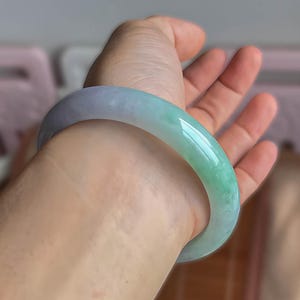 Puede incluir: Un brazalete de jade en tonos verdes y lavanda. La piedra pulida es lisa y curvada, con un degradado de colores. El brazalete se lleva en la muñeca, mostrando su belleza natural y diseño.