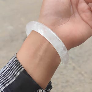 Może przedstawiać: Przezroczysta biała bransoletka typu bangle. Bransoletka ma gładki, zaokrąglony kształt z lekko nieregularną powierzchnią. Światło przechodzi przez bransoletkę, nadając jej miękki, świecący wygląd. Bransoletka jest noszona na nadgarstku, a widoczny jest rękaw czarno-białej koszuli w kratę.