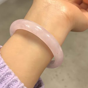 Op de afbeelding: Een lichtroze jade armband. De gladde, afgeronde armband wordt om een pols gedragen, met een lichtpaarse trui zichtbaar. De armband is zacht, doorschijnend roze.