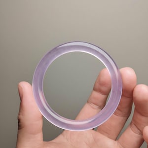 May include: A translucent lavender-colored jade bangle bracelet. The smooth, polished surface reflects light, showcasing the natural variations in the stone. The bracelet is a complete circle, designed to be worn on the wrist.