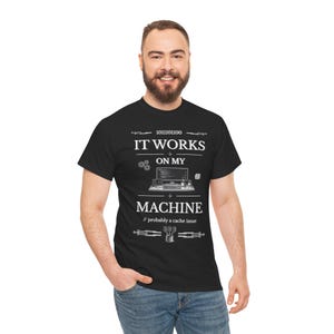 Op de afbeelding: Een zwart t-shirt met korte mouwen en witte grafische tekst met de tekst IT WORKS ON MY MACHINE en // probably a cache issue. Het ontwerp bevat een binaire codereeks, een illustratie van een computerwerkstation en kleine tandwiel- en databasepictogrammen.