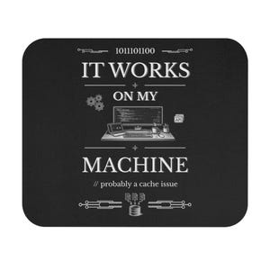 May include: A rectangular black mouse pad featuring white text that reads IT WORKS ON MY MACHINE, with a graphic of a computer setup, binary code, gears, and the text // probably a cache issue, centered on the surface with decorative line accents.