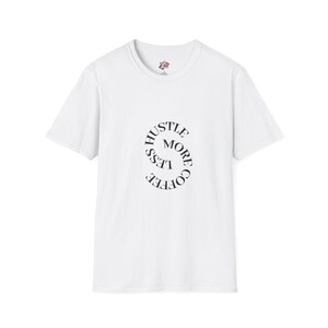May include: White t-shirt with the text "Hustle More, Coffee Less" in a curved, circular design. The shirt has a crew neck and short sleeves. The text is in a dark, sans-serif font.